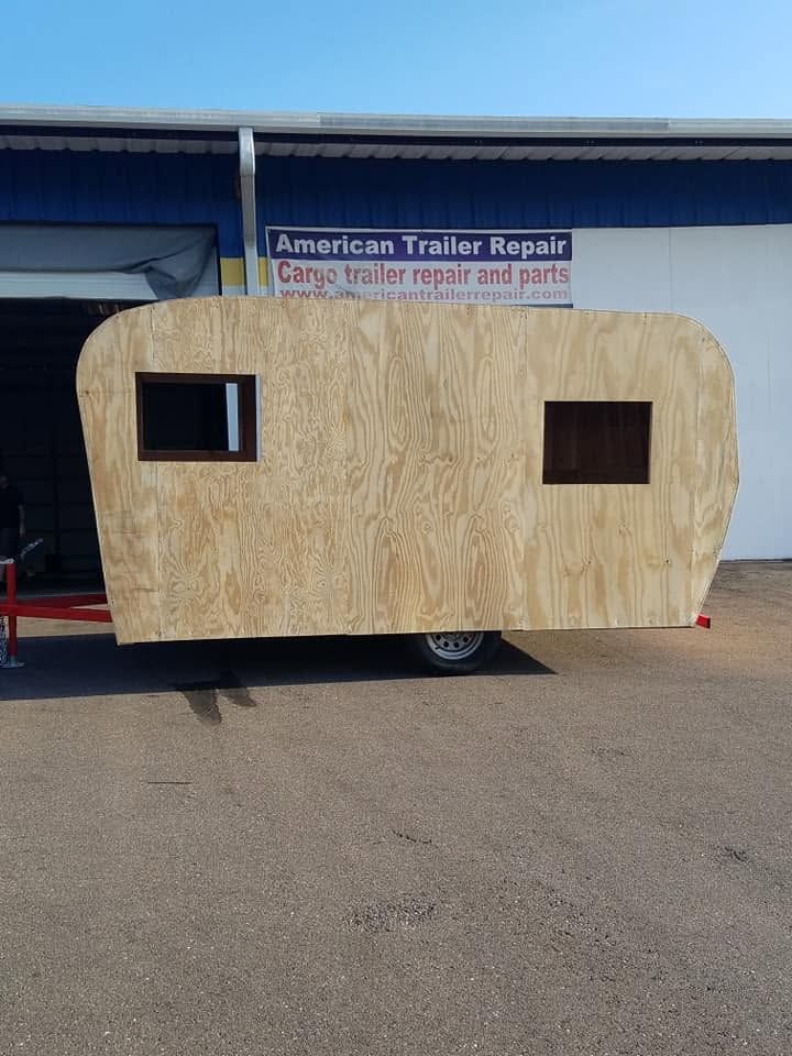 Wood-paneled trailer with two small windows parked outside a repair shop.