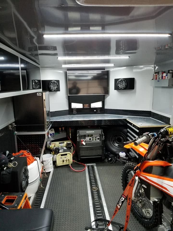 Inside a trailer garage, orange dirt bikes and tools are arranged under bright ceiling lights.