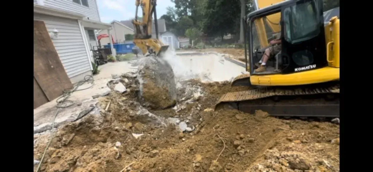 Excavator lifting a large boulder in a muddy residential driveway beside a house