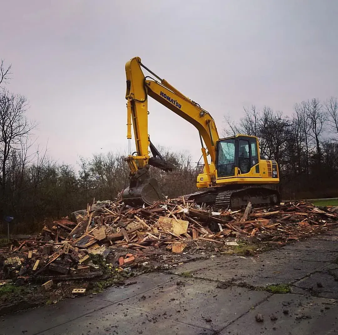 Yellow excavator clearing debris in a muddy lot under an overcast sky