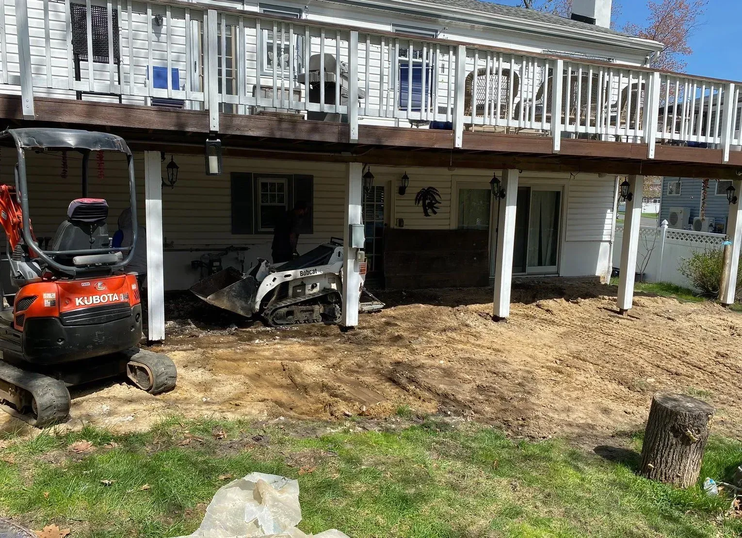 Orange lawn tractor and white skid steer under a raised house deck with wooden posts and grass below