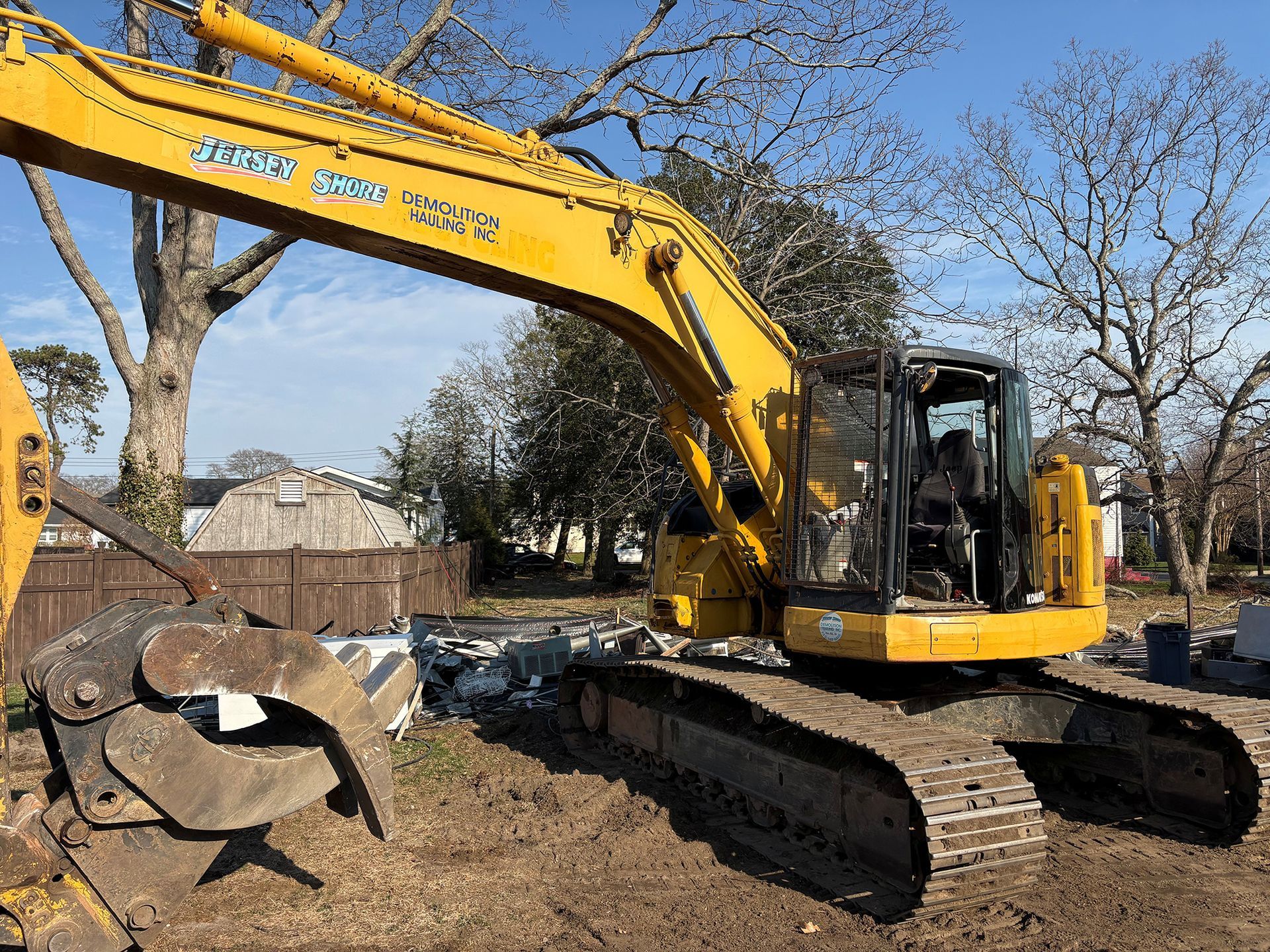 Yellow excavator on tracks beside a large tree stump in a yard with a wooden fence.