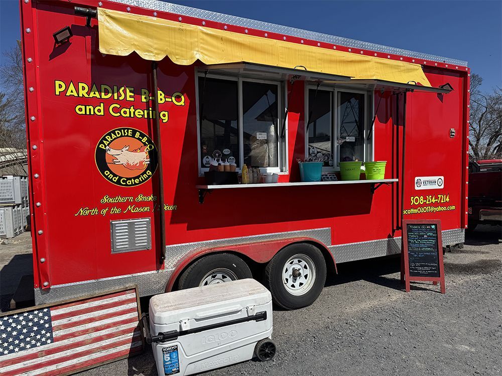 Red food truck with serving window and menu sign, parked on gravel outdoors