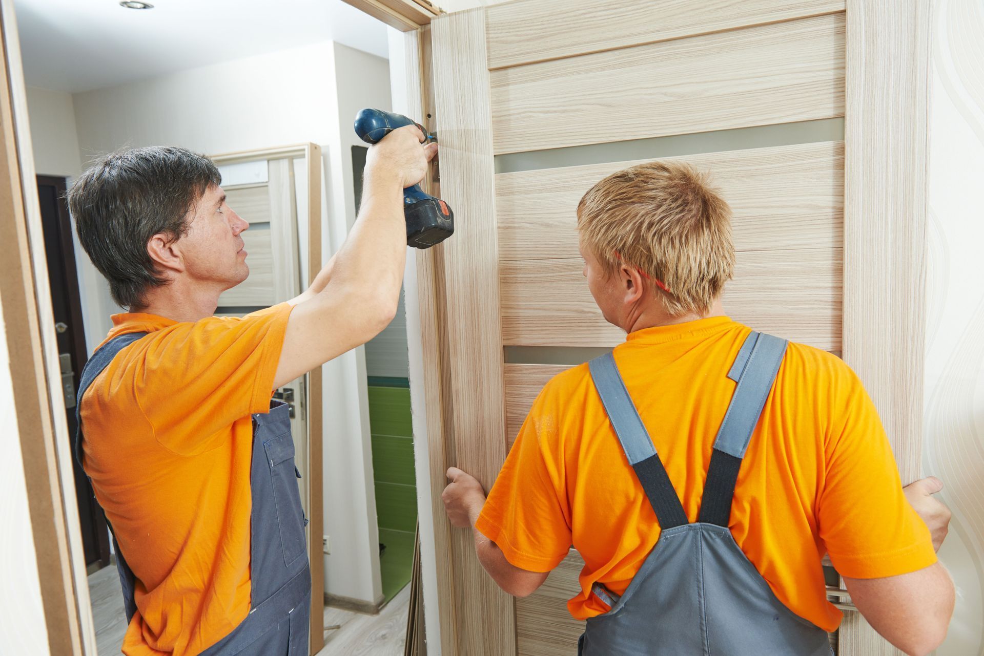 Two construction workers installing a door frame, one using a drill, wearing orange shirts and overalls.
