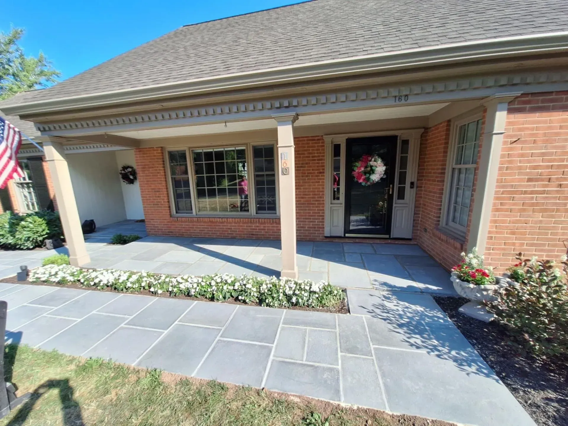 Exterior view of a brick house with a covered porch and blue stone walkway. The front door has a wreath and is flanked by windows and flower pots.