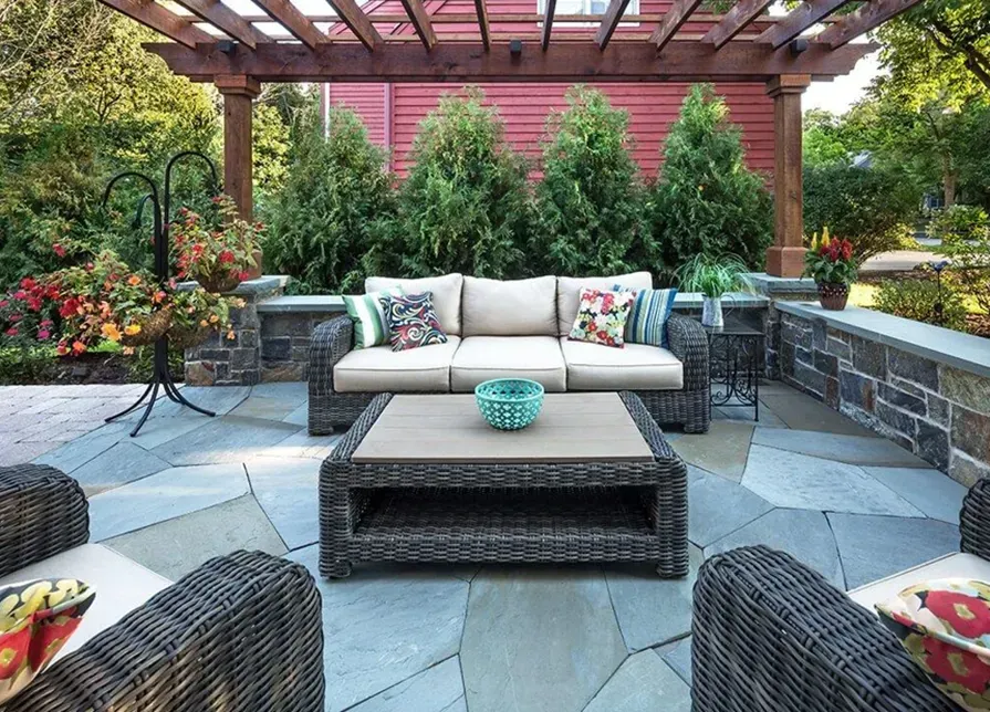 Outdoor patio with wicker furniture, a stone floor, and a pergola. The background features evergreen trees and a red wall.