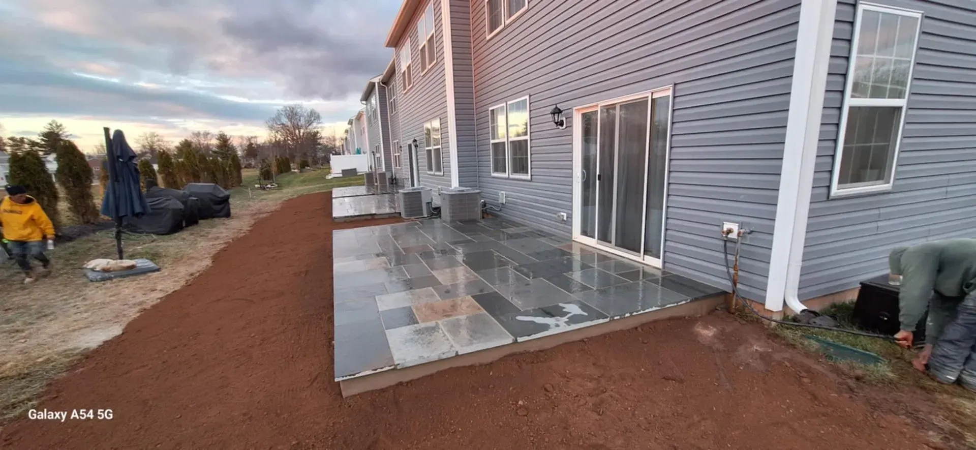A house with a gray siding, a paved patio in front of a sliding glass door, and two people working on the ground.