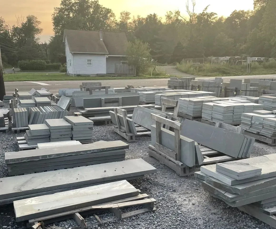 Stone slabs and pavers stacked on pallets in an outdoor lot; a house and trees are in the background.