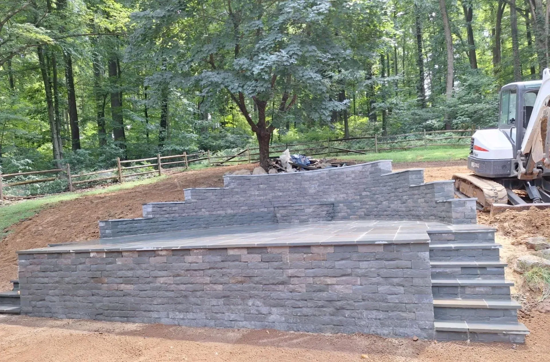 A tiered outdoor stage built with gray stone blocks; brown dirt surrounds it. A small excavator sits to the side, woods in the background.