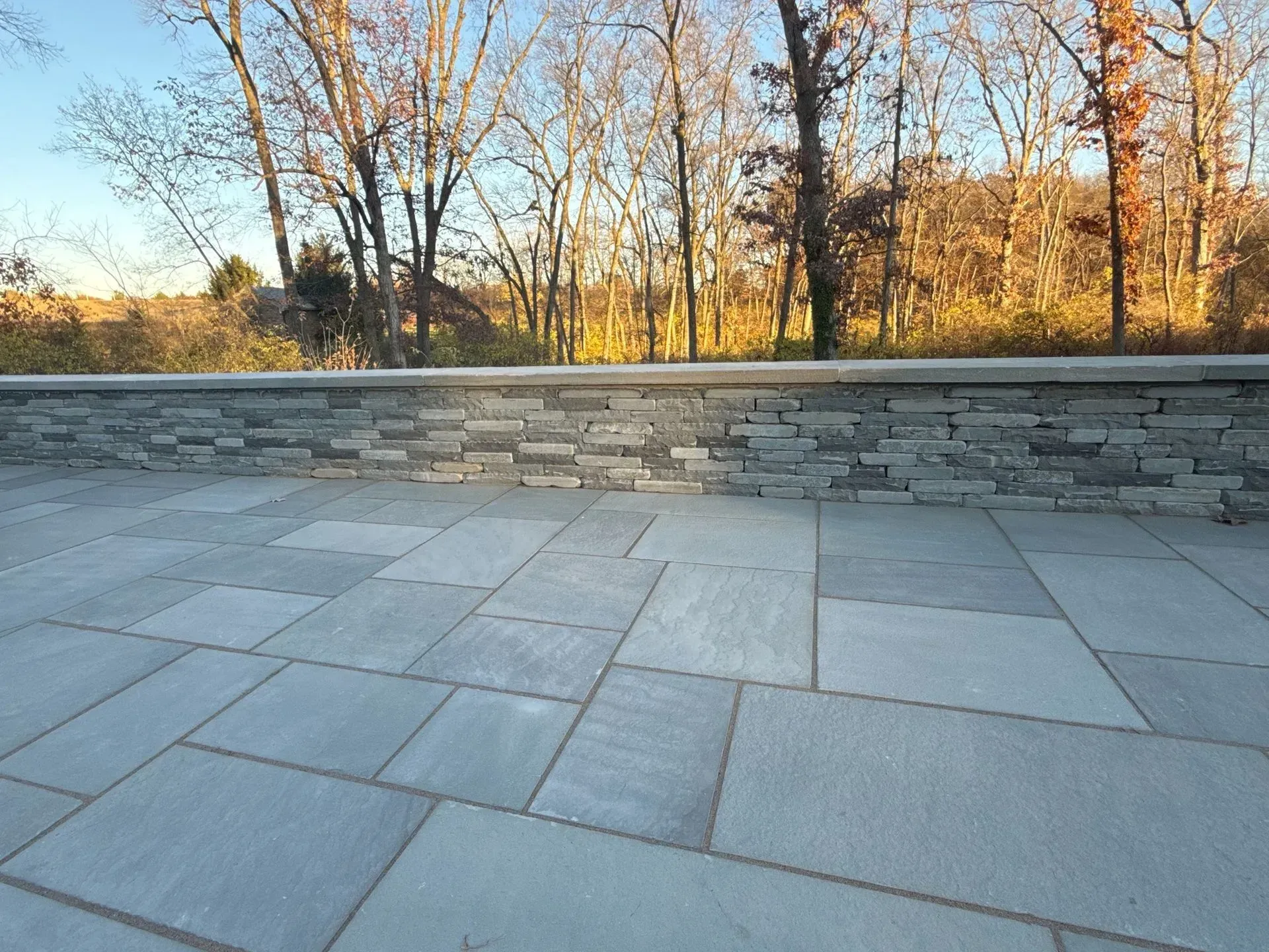 A stone patio with a stacked stone wall in front of a wooded area during daytime. The patio tiles are blue-gray.