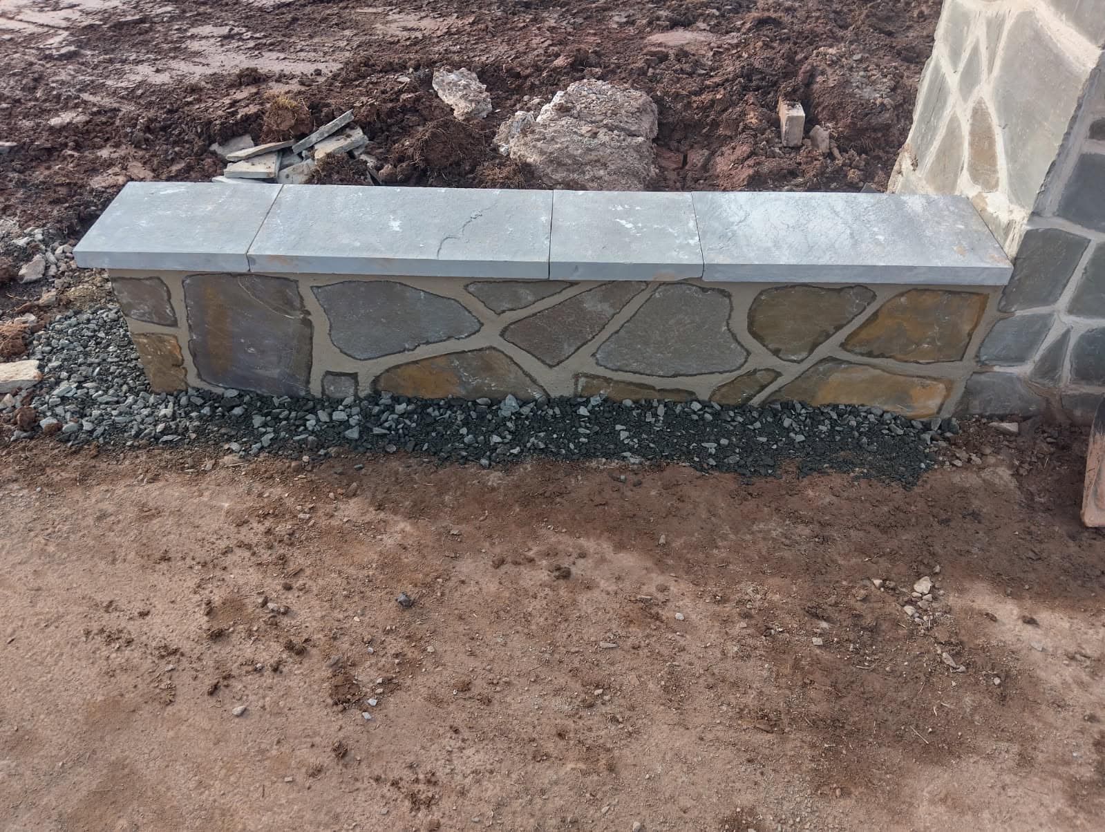 A stone bench with a gray stone top and a mosaic-style stone front, set on gravel. Brown dirt surrounds it.