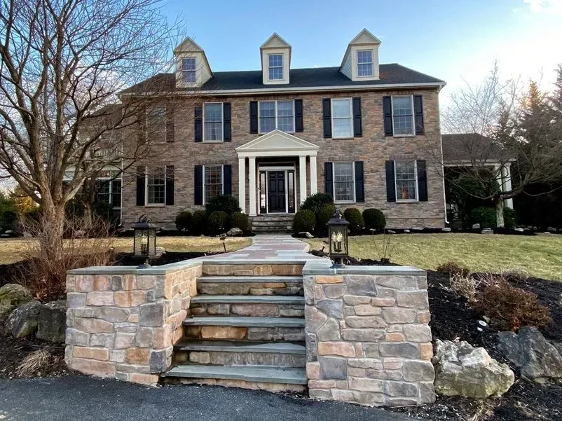 Two-story brick house with stone steps and a walkway. Black shutters, a white portico, and lanterns flank the entrance.