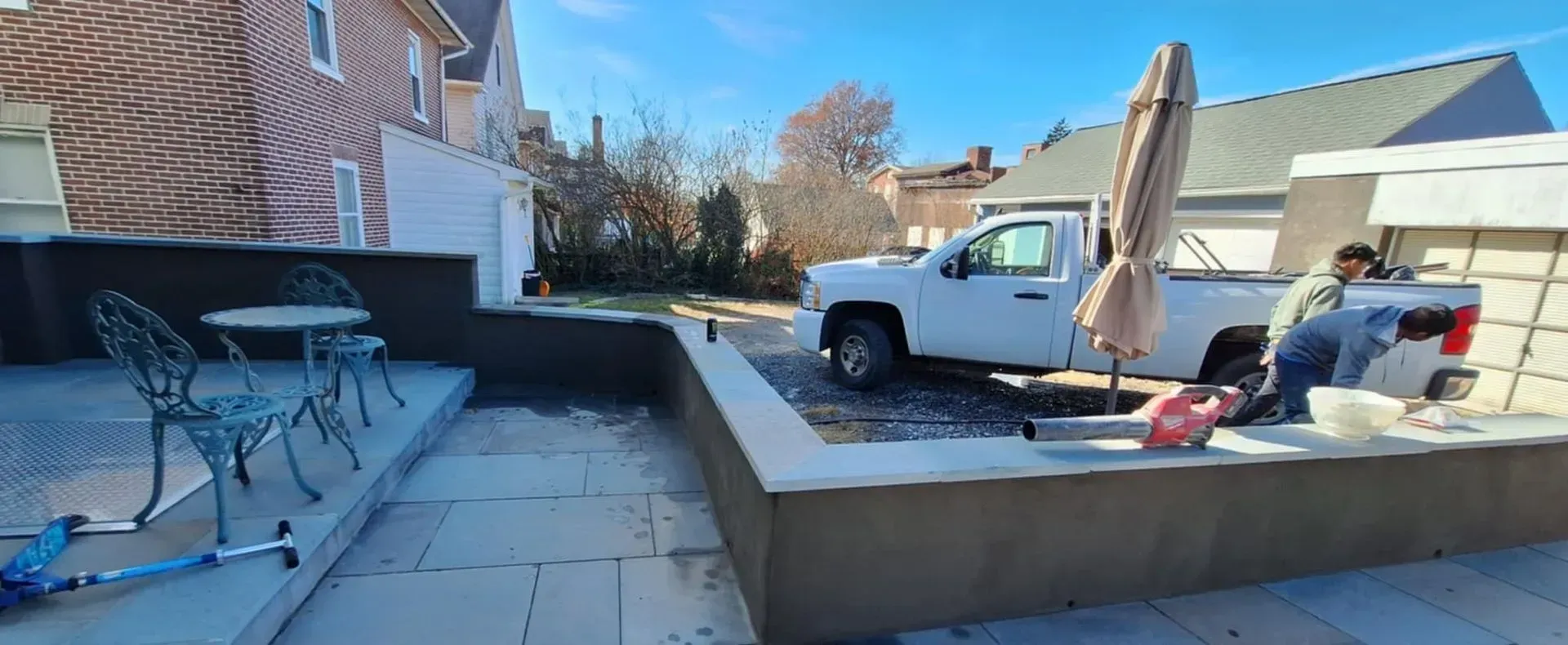 Backyard with a white truck, two people working, and patio furniture. Red brick house and blue sky in the background.