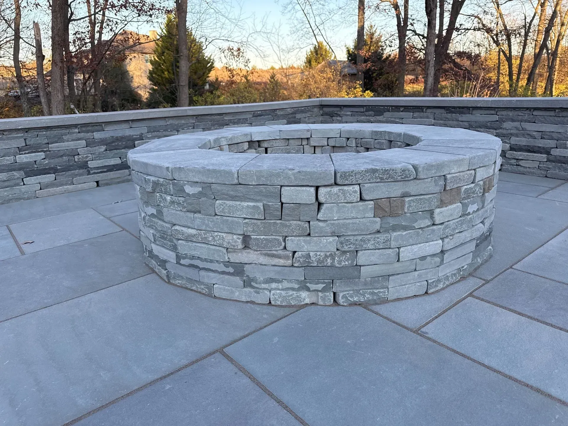 A round stone fire pit on a gray stone patio. Low stone walls surround the patio, with trees and a building in the background.