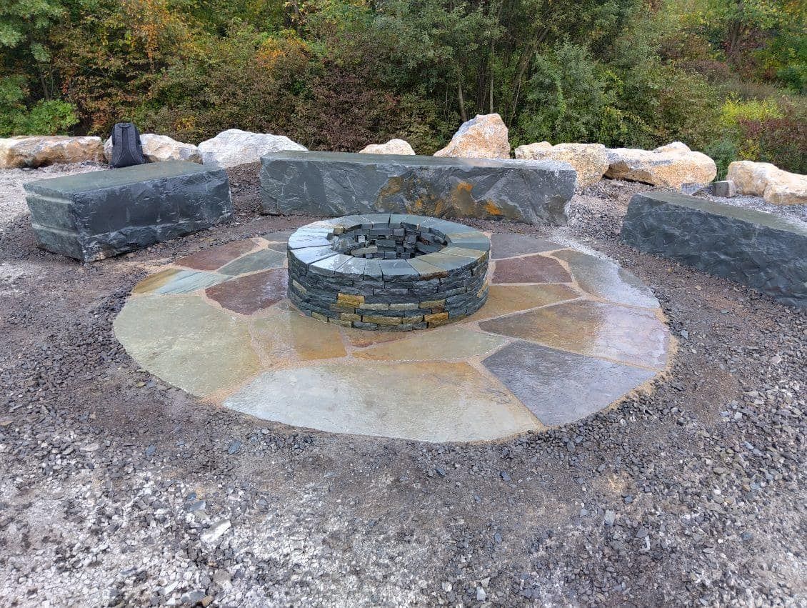 Stone fire pit surrounded by flagstones and seating benches in a natural outdoor setting.