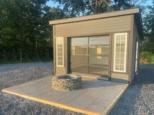 Small gray shed with a glass garage door, white window frames, and a stone fire pit on a concrete patio.