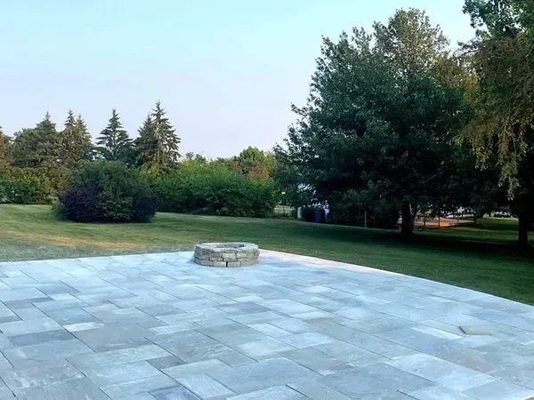 A stone patio with a fire pit in the center, surrounded by a green lawn and trees under a pale blue sky.