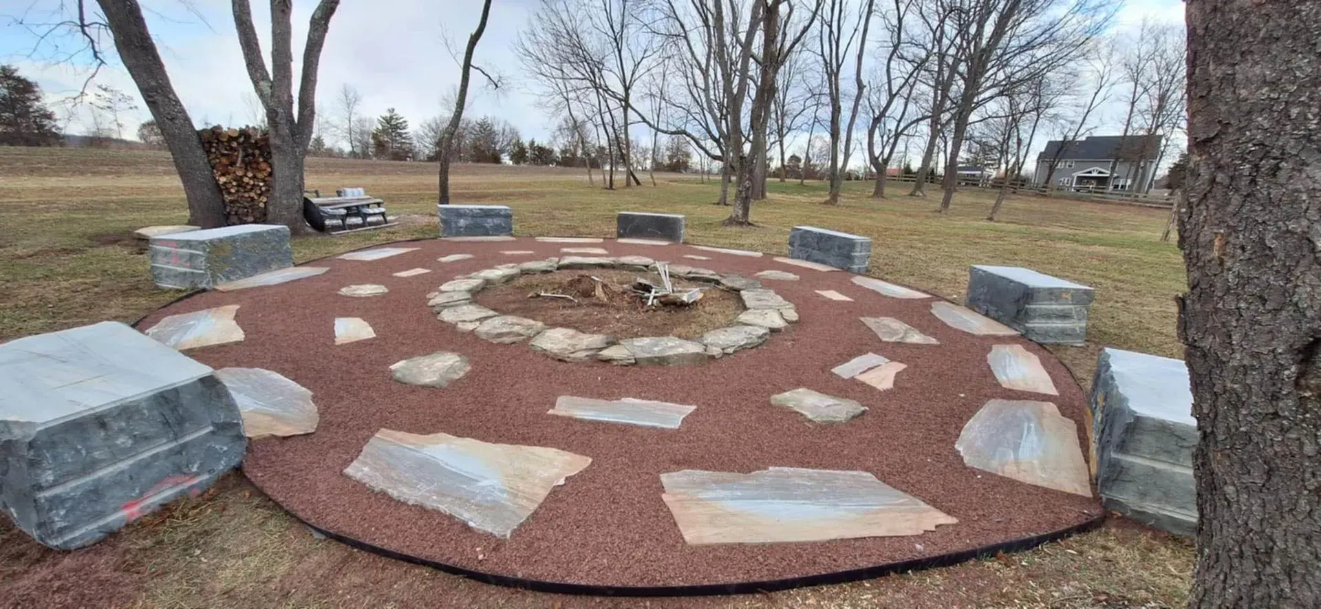 A circular fire pit area with large stone seating and red gravel in a grassy yard, trees in the background.