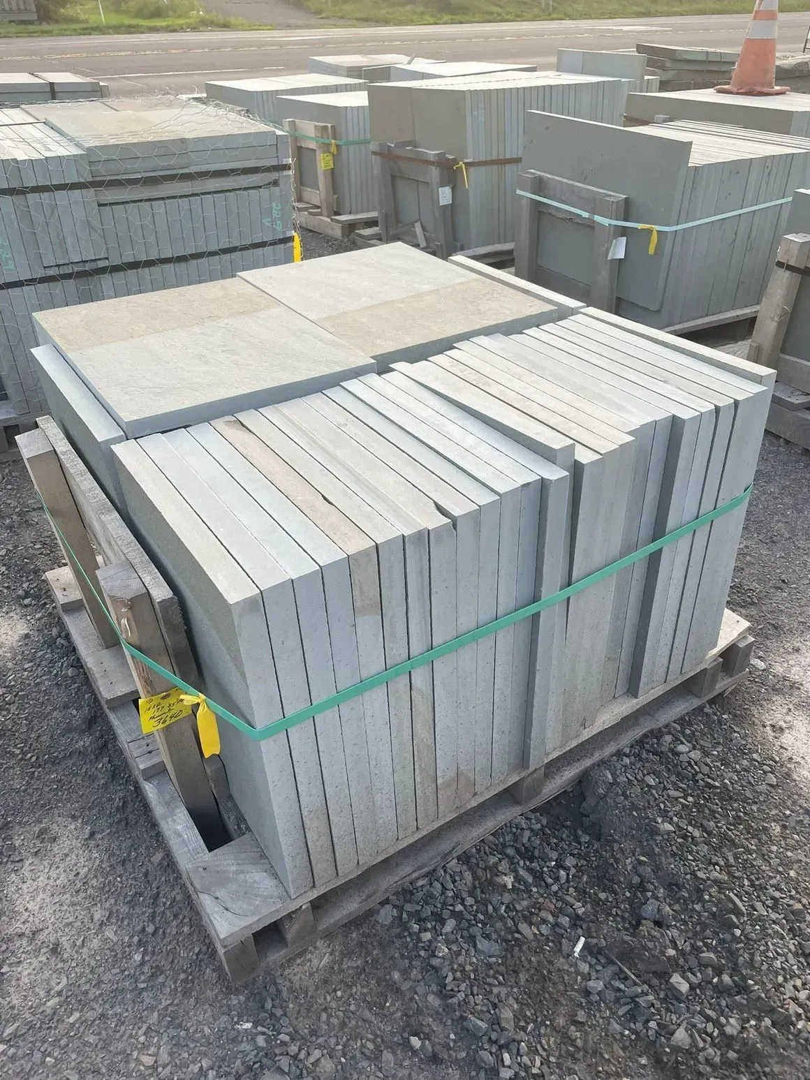 Stacks of gray stone pavers on wooden pallets, secured with green straps, outdoors on gravel.