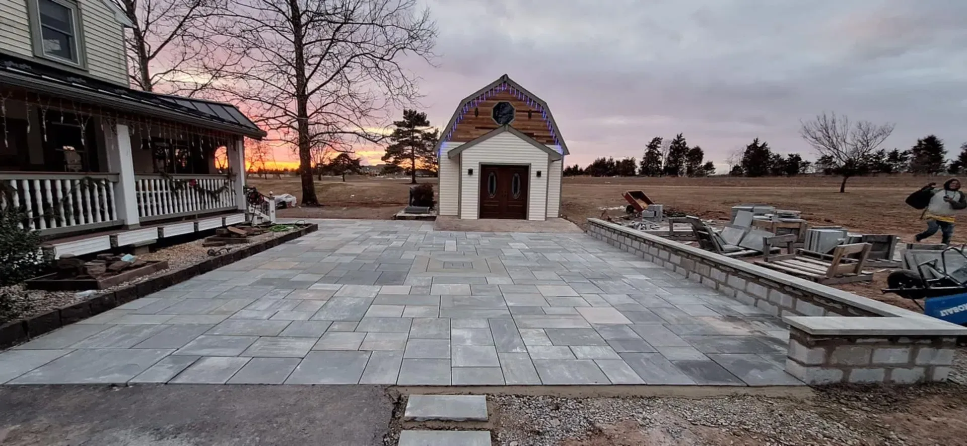 A house with a porch and a small barn-shaped building, connected by a paved patio, set against a colorful sunset.