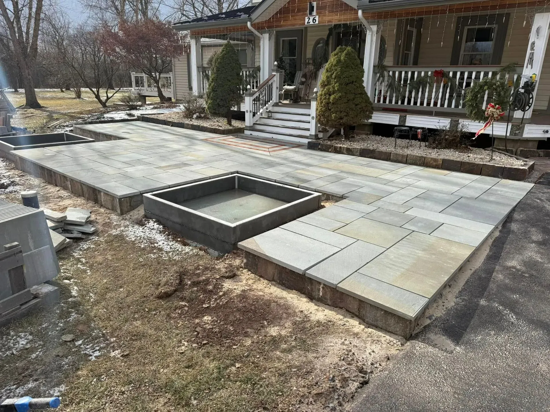 Exterior view of a house with new stone patio and a square, raised planting bed. Construction debris is visible around the edges.