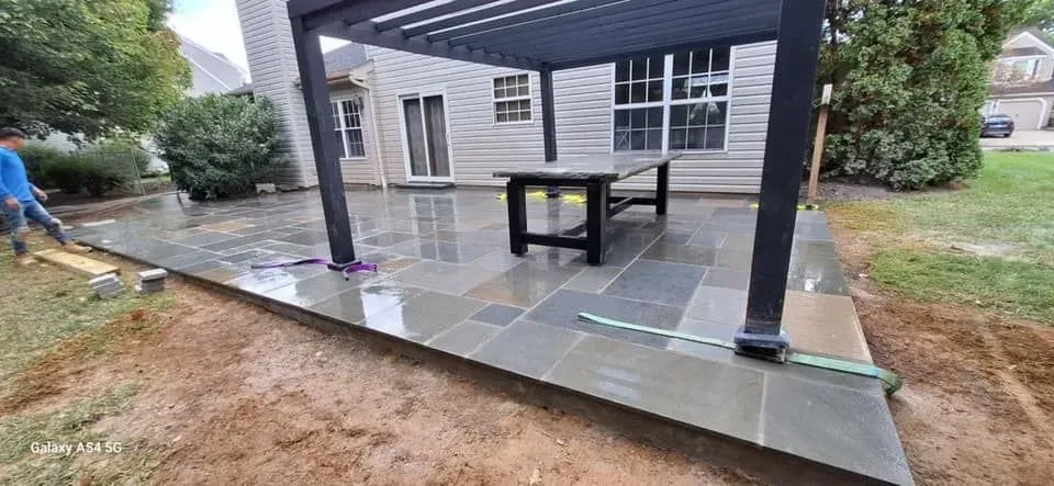A newly constructed patio with stone tiles and a pergola with a table, next to a house and lawn.