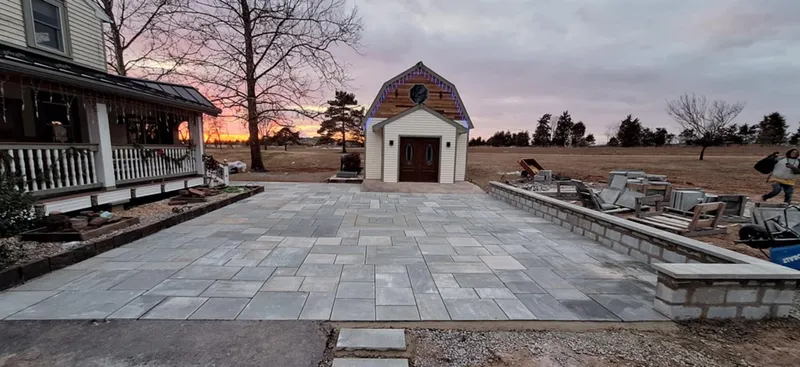 Newly constructed stone patio with a small building and house in the background at dusk.