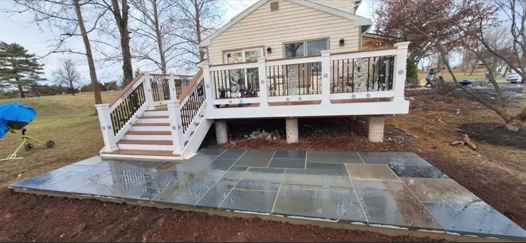 A backyard patio with a deck, steps, and flagstone. The deck has white railings and dark decorative details, the patio and steps are wet.
