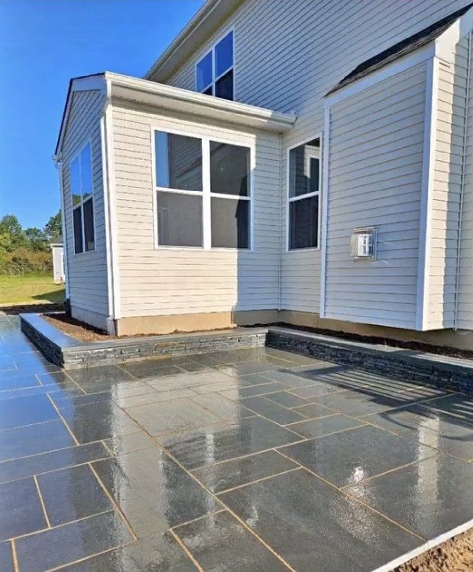 Gray stone patio next to a light-colored house with windows on a sunny day.