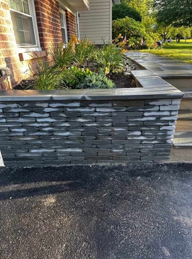 Stone retaining wall with plants, adjacent to a paved driveway and steps leading to a brick house.