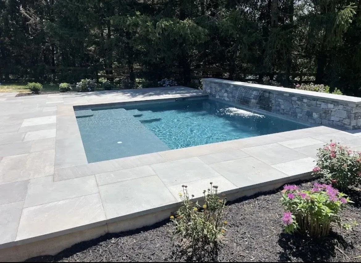 Rectangular backyard pool with stone surround and small waterfall feature. Gray stone patio, dark mulch, and blooming pink flowers.