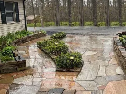 Flagstone patio with rectangular stone planters filled with green bushes. A wet driveway leads to the trees.