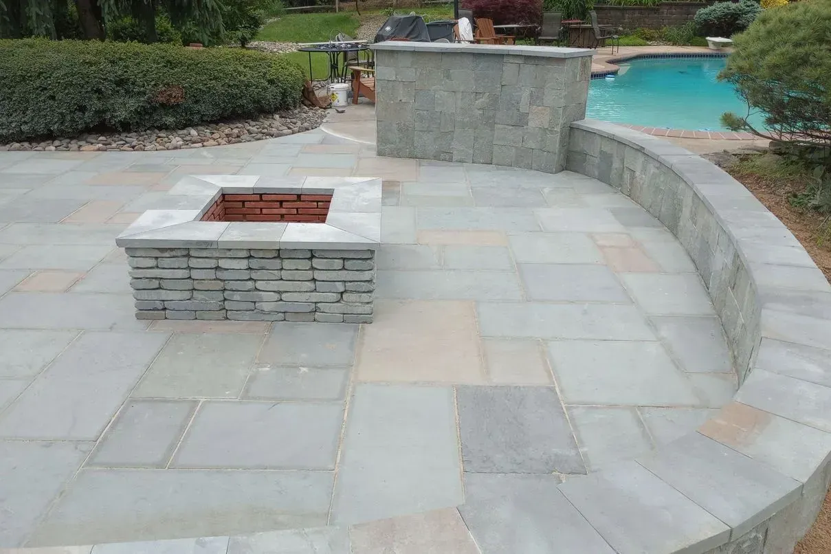 Stone patio with built-in fire pit and bar next to a swimming pool. Green and gray stone, with blue water in the background.