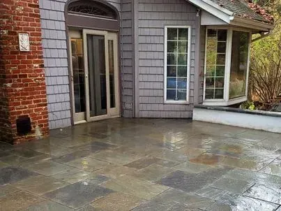 Exterior patio with wet, dark stone tiles next to a house with gray shingles, a brick chimney, and a bay window.
