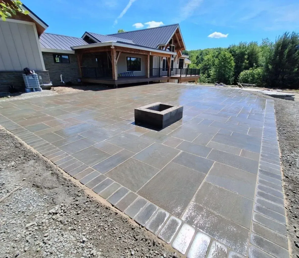 Newly constructed stone patio with a fire pit, bordering a house with a wooden porch under a blue sky.