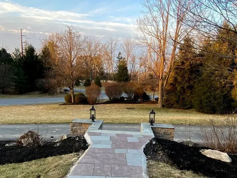 Brick walkway leading to a home with lanterns, trees, and shrubs in the yard. Overcast sky.