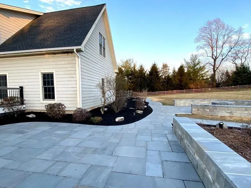 Backyard patio with blue stone pavers, a gray retaining wall, and a light-colored house with a dark roof.