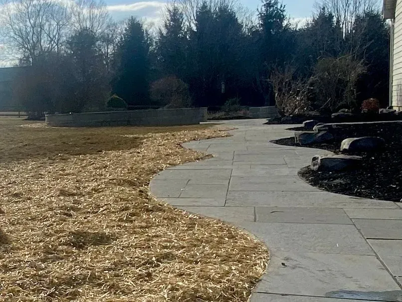 A stone pathway curves through a yard with wood chips and landscaping. Trees line the background under a sunny sky.