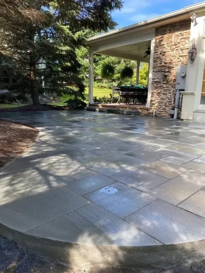 A newly constructed gray stone patio with a curved edge leads towards a covered porch with a stone wall.