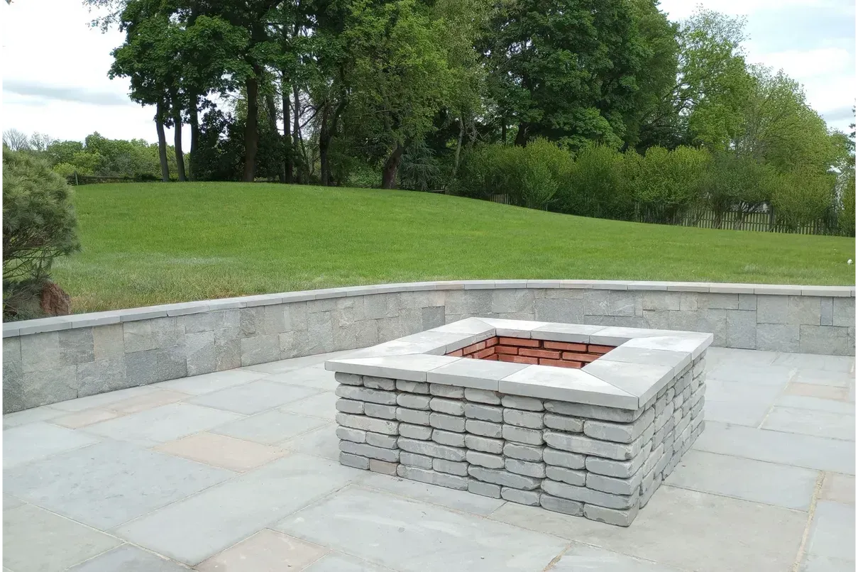 A stone fire pit on a paved patio, with a grassy hill and trees in the background.
