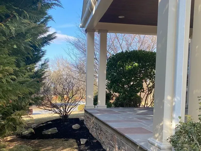 A covered porch with white columns and stone flooring. Trees and bushes surround the porch, with a blue sky visible.