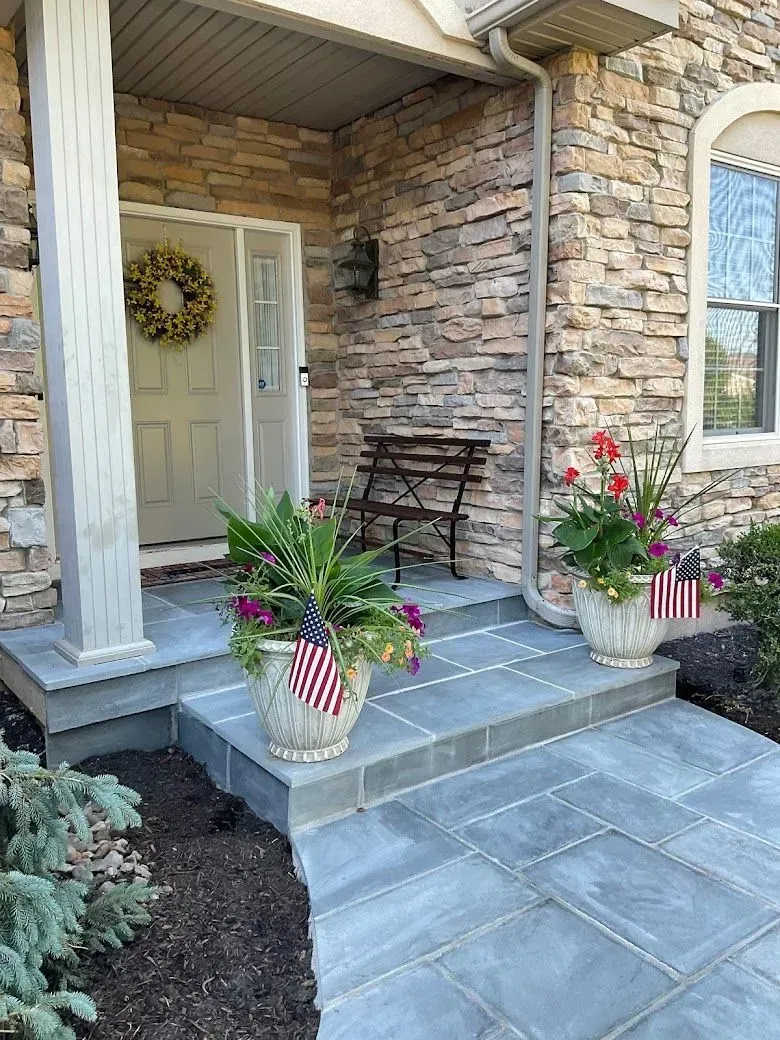 Stone entryway of a house with blue flagstone steps. Potted plants with American flags flank the door, with a bench and wreath.