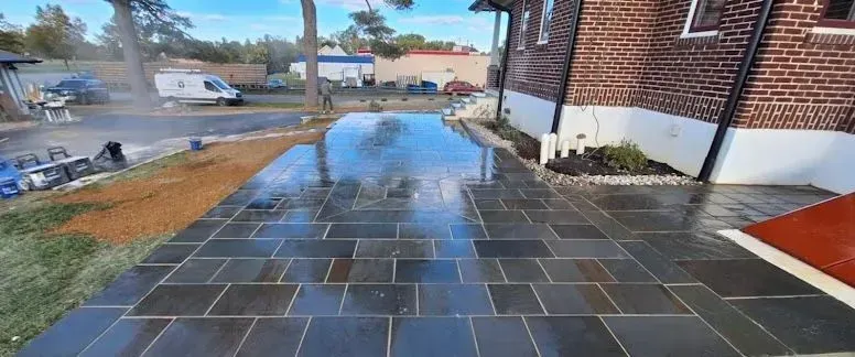 Exterior view of a newly paved patio with wet, dark gray pavers. A brick building and grassy area are to the right and left.