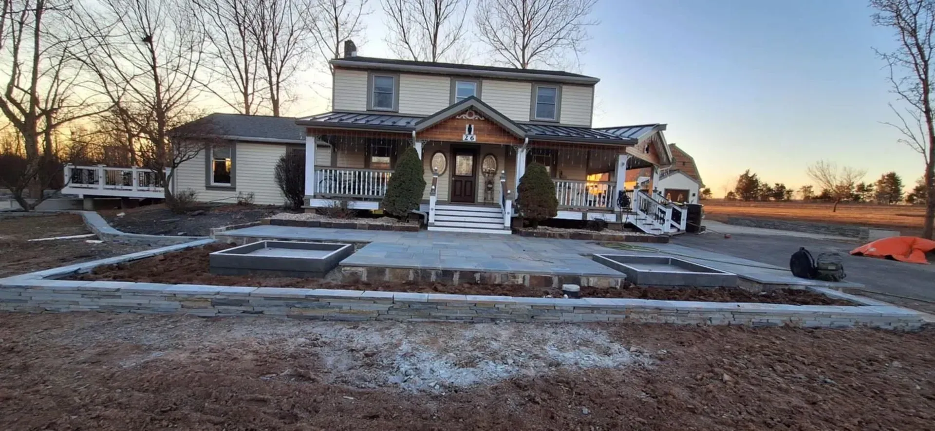 A two-story house with a porch is under construction. A concrete patio and garden beds are in the foreground. The sky is clear.