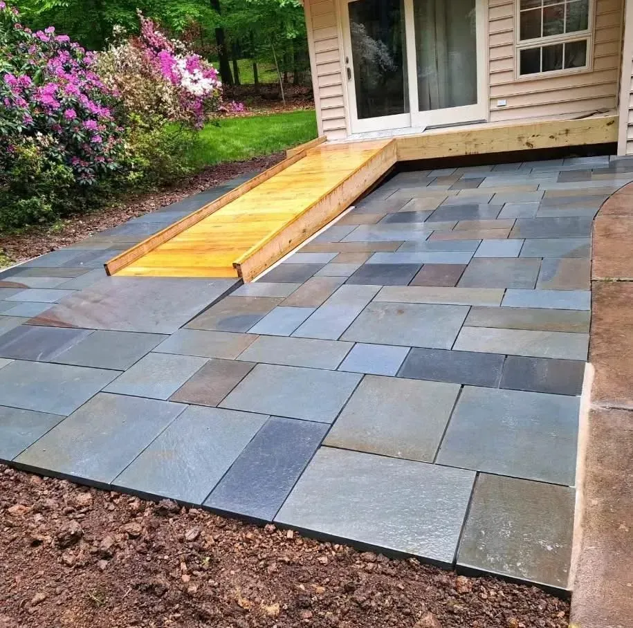 Blue stone patio with a wooden ramp leading to a light-colored building. Green grass and pink flowers are in the background.