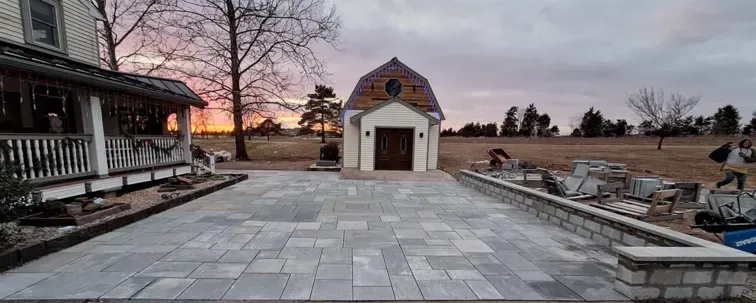 A paved patio with a small building, a house, and a setting sun in the background. The sky is cloudy.