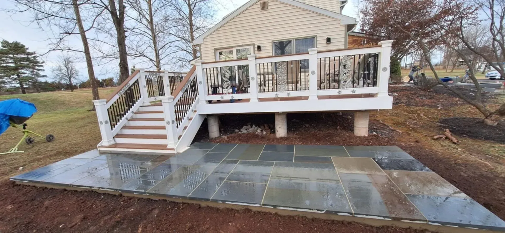A stone patio in front of a deck with white railings and stairs. The house is beige, and the ground is brown with trees in the background.