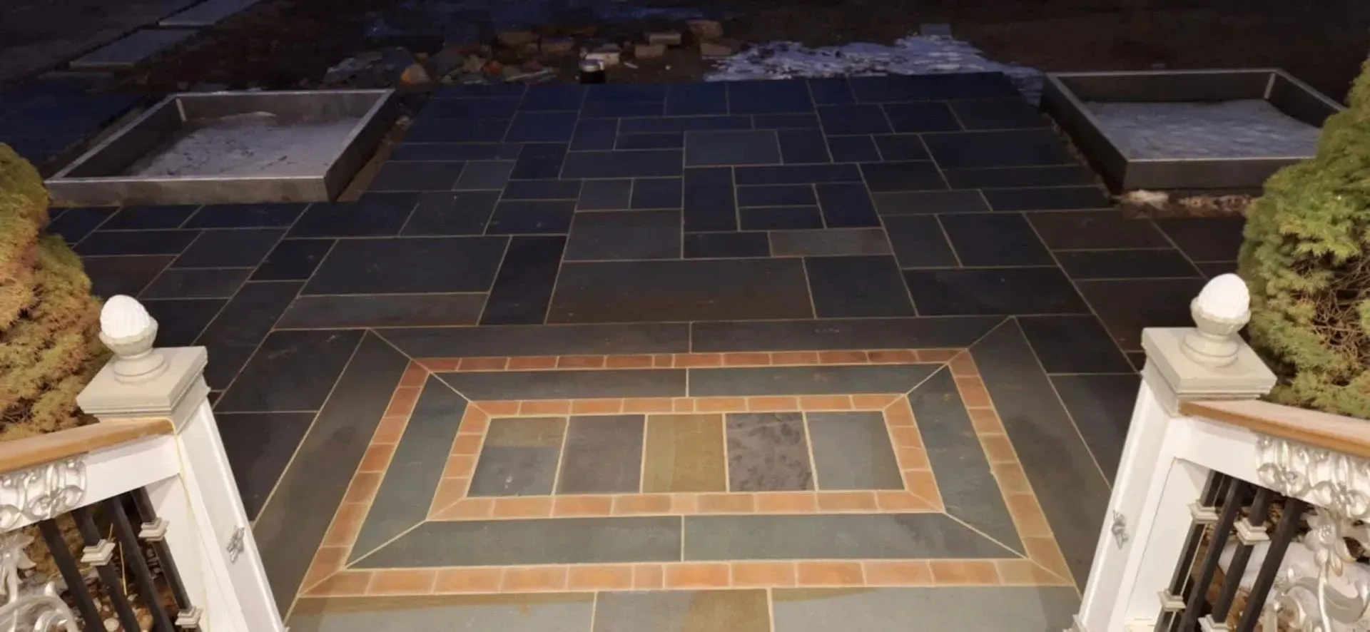 View from top of stairs of a brick patterned walkway with decorative tile design. Two square planters are positioned on either side.