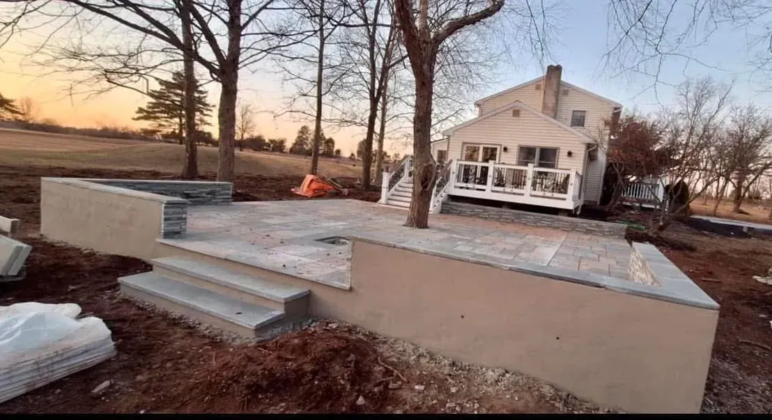 Outdoor patio construction with concrete steps and retaining walls next to a two-story house and trees.