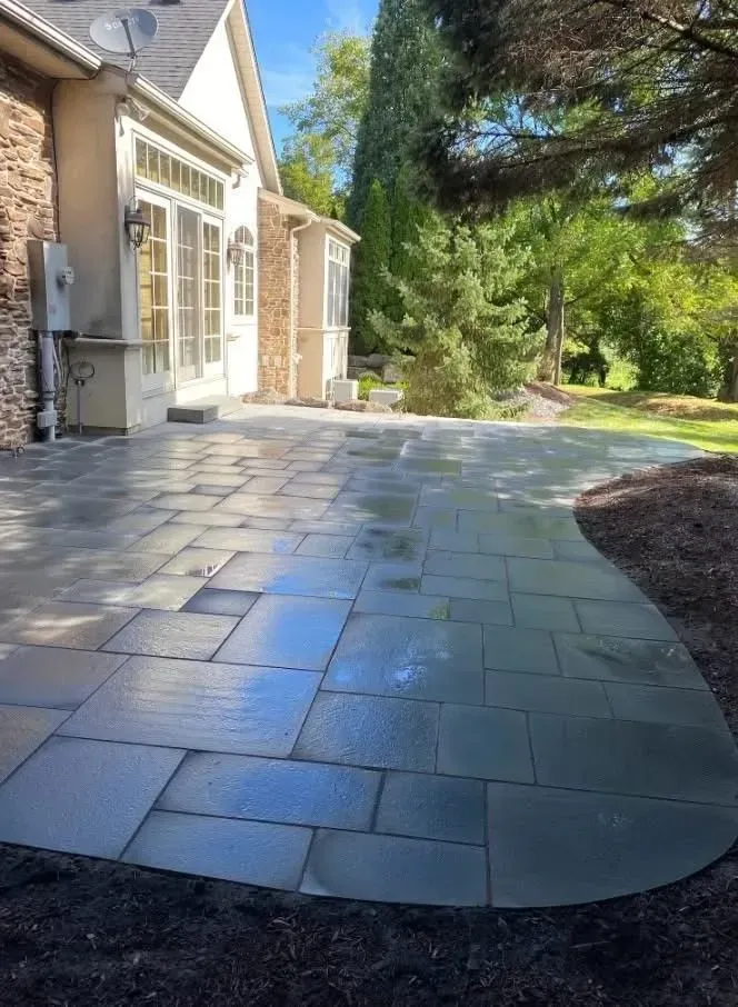 Gray stone patio extending from a light-colored house, with lush green grass and trees in the background.